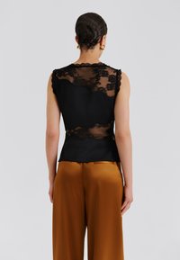 Top negru fără mâneci cu spate din dantelă cu modele florale și margini ondulate, asortat cu pantaloni din satin maro deschis.