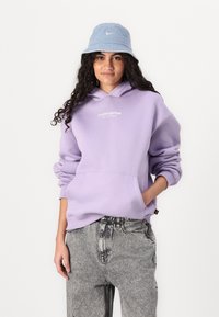 Lilla hettegenser med teksten "STUDIO EDITION", frontlomme og ribbestrikkede mansjetter, kombinert med grå mønstrede baggy jeans og en lyseblå bøttehatt.