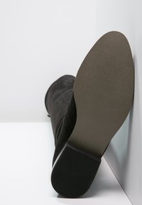 Bota de ante negra hasta la rodilla con un eje texturizado y una suela de goma. La parte inferior cuenta con un dibujo en la suela para mayor agarre.