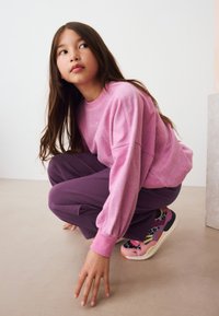 Růžová mikina s dlouhými rukávy, doplněná fialovými tepláky. Barevné tenisky s různorodými texturami a vzory. Jednoduché pozadí.