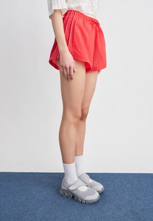 Personne portant un short de sport rouge avec des bandes blanches, des chaussettes blanches et des baskets en mesh gris, debout sur un tapis bleu contre un mur blanc.