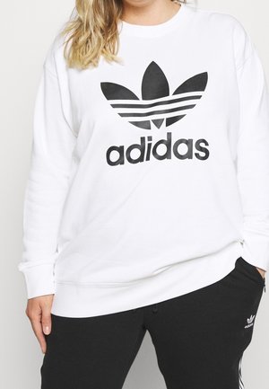 Person iført hvid Adidas-sweatshirt med sort logo og sorte Adidas-bukser, stående med den ene hånd i lommen og den anden ned langs siden.