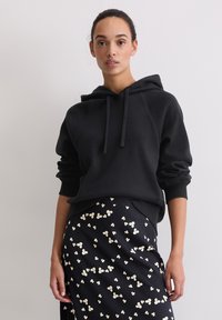 Felpa nera con cappuccio e vestibilità rilassata, con polsini e orlo a coste. Abbinata a una gonna scura decorata con piccoli motivi floreali.