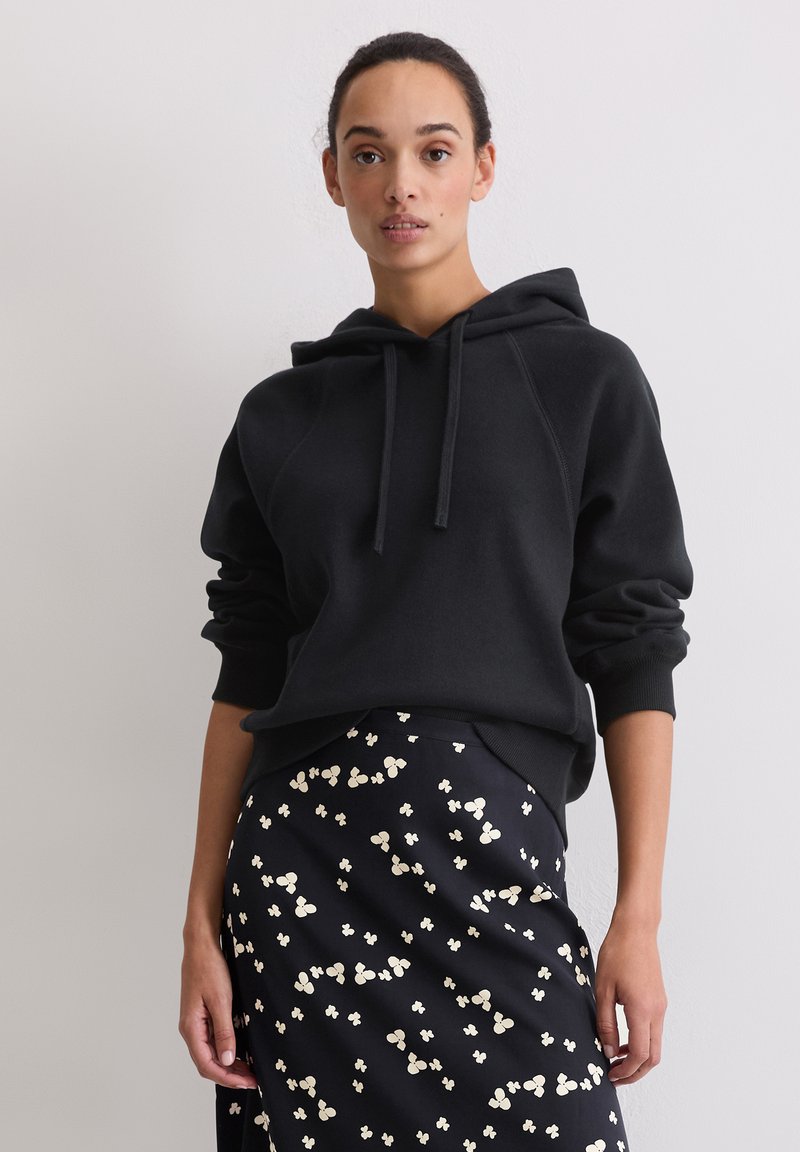 Felpa nera con cappuccio e vestibilità rilassata, con polsini e orlo a coste. Abbinata a una gonna scura decorata con piccoli motivi floreali.
