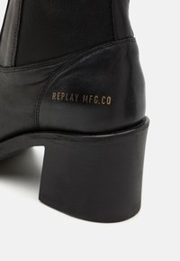 Crne kožne čizme do gležnja s teksturiranim elastičnim umetkom, masivnim potplatom i zlatno utisnutim logotipom "REPLAY MFG.CO" na stražnjem dijelu.