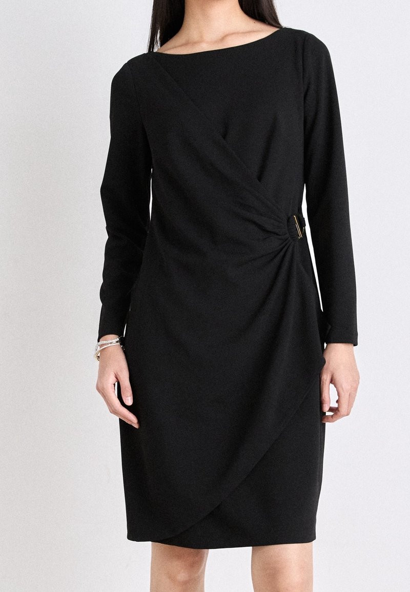 Femme portant une robe noire à manches longues avec un détail de boucle latérale et un tissu froncé à la taille, debout devant un fond clair.