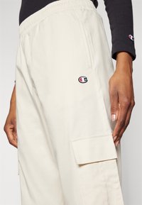 Béžové cargo kalhoty s volným střihem, které mají elastický pas a boční kapsy. Na stehně se nachází malá černá a červená nášivka.