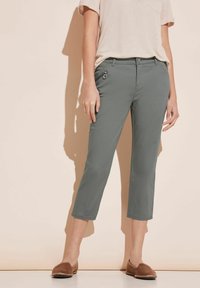 Nedre delen av en person som bär grå kapade byxor med dragkedjefickor, en beige kortärmad topp och bruna slip-on skor mot en beige vägg.