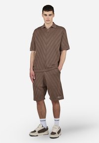 Brun strikket polo og shorts-sett med et chevronmønster. Har krage, ribbede mansjetter og logo detalj på shortsen. Hvite sneakers på føttene.