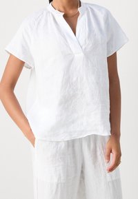 Haut blanc en lin à manches courtes avec un col montant, une coupe décontractée et une surface légèrement texturée, porté avec un pantalon blanc ample.