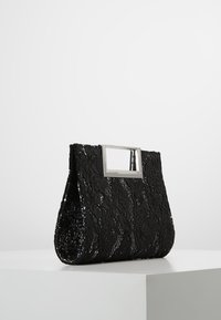 Mustaa pitsiä oleva clutch-laukku kukkakuviolla ja suorakulmaisella hopeakenkällä, seisoo valkoisella jalustalla vaaleanharmaata taustaa vasten.