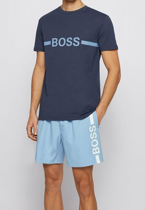 Muž ve tmavě modrém tričku s nápisem "BOSS" a světlých modrých šortkách s vertikálním nápisem "BOSS", stojící s jednou rukou v kapse.