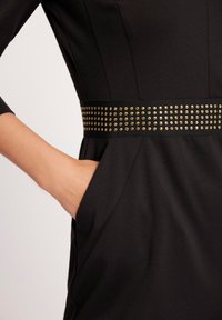 Czarna sukienka o dopasowanym kroju, z rękawami 3/4 oraz szerokim czarnym paskiem ozdobionym złotymi ćwiekami. Kieszeń widoczna w szwie bocznym.