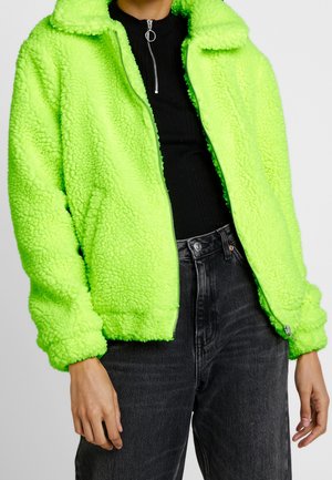 Person trägt eine leuchtend neongrüne, flauschige Jacke über einem schwarzen Zip-up-Oberteil und dunkelgrauen Jeans und steht vor einem schlichten Hintergrund.