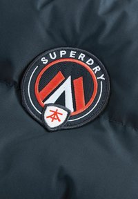 Čierna tkanina s kruhovou vyšitou nášivkou s červenými a bielymi akcentmi. Text obsahuje "SUPERDRY" a stylizované červené logo vo vnútri.