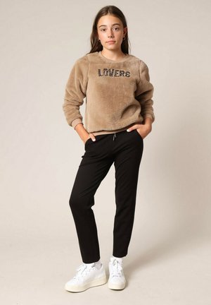 Tan fleece sweater met "LOVERS" in zwart, gecombineerd met zwarte taps toelopende broeken en witte sneakers. De stof is zacht en heeft een relaxte pasvorm.
