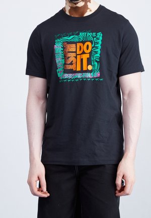 Mężczyzna w czarnej koszulce z pomarańczowym, zielonym i różowym napisem "JUST DO IT." oraz wieloma mniejszymi logo "Nike" na przodzie.