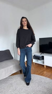 Γυναίκα με μακριά καστανά μαλλιά, φοράει μαύρο πουλόβερ και μπλε τζιν, στέκεται άνετα σε ένα σαλόνι με καναπέ, τηλεόραση και λευκή ντουλάπα.