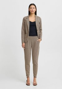 Bruine tweed blazer met bijpassende taps toelopende broek, gecombineerd met een zwart topje. Zachte textuur, blazer met enkele knoopsluiting, broek tot de enkel.