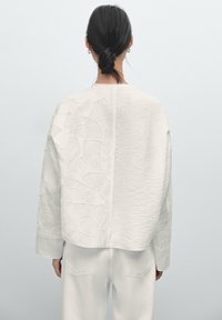 Donna con capelli scuri raccolti in un basso chignon, indossa un top bianco a maniche lunghe testurizzato e pantaloni bianchi, vista di spalle su uno sfondo neutro.