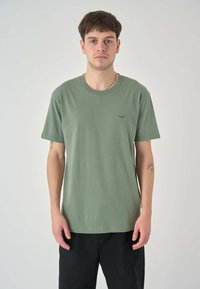 Grünes kurzärmliges T-Shirt aus weichem Stoff, mit rundem Ausschnitt und einem kleinen dunkelgrünen Logo auf der linken Brust.