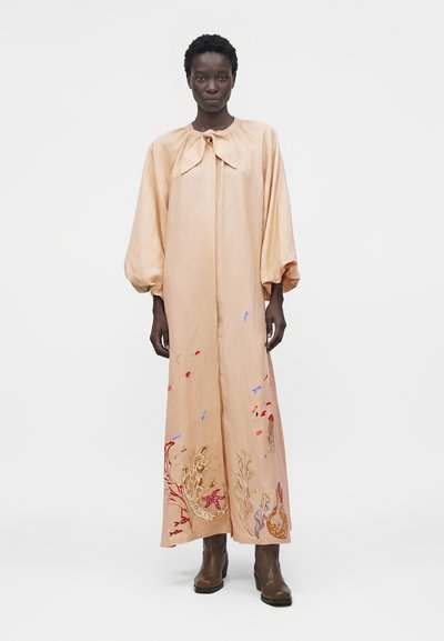 Γυναίκα που φοράει ένα μακρύ ροδακινί φόρεμα με μπαλόν μανίκια, λαιμόκοψη με φιόγκο, κεντητά σχέδια με θαλασσινά θέματα και καφέ μποτάκια στο ύψος των αστραγάλων, σε προσκήνιο.