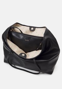 Sort læder tote taske med en tekstureret overflade, indeholdende et beige interiør med et gentagende "GUESS" mønster og en lynlåslomme.