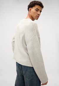 Lys grå strikkegenser med løs passform, senkede skuldre og rund hals, kombinert med mørkeblå jeans. Myk tekstur, uformell stil.