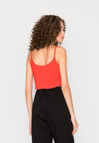 Top cropped vermelho com nervuras, com alças finas e uma bainha com babados, combinado com calças pretas de cintura alta, mostrado de lado.