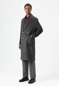 Grauer doppelt geknöpfter Wollmantel mit großen Knöpfen, aufgesetzten Taschen und lockerer Passform. Kombiniert mit grauer Hose und schwarzen Schuhen.