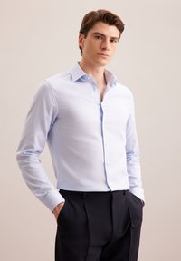 Jasnoniebieska koszula z długim rękawem z guzikami i rozkładanym kołnierzykiem, wykonana z gładkiego materiału. Noszona z ciemnymi spodniami, ręce włożone w kieszenie.