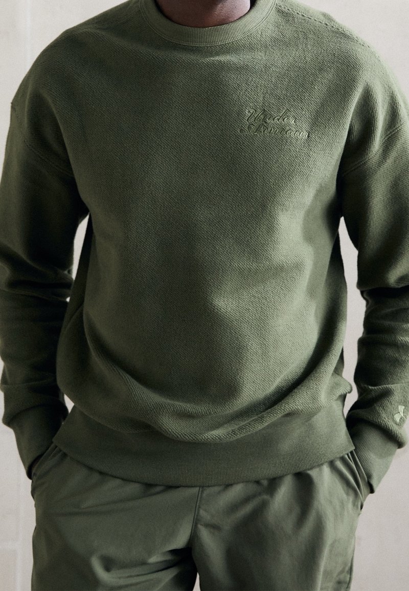 Homme portant un sweat-shirt Under Armour vert olive et un pantalon assorti, les mains dans les poches, debout devant un fond neutre.