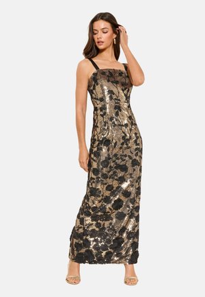 Naine kuldse litrite ja mustade lillemustriga varrukateta kleidis, millel on mustad õlapaelad ja beežid avatud varbaga kontsakingad, poseerimas ühe käega juuste juures.