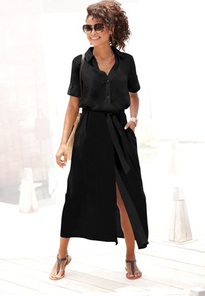 Woman wearing sunglasses and a black short-sleeve button-up top with a matching long skirt with a front slit, sandals, and carrying a woven bag.