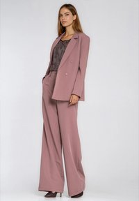 Dopasowana marynarka dwurzędowa i spodnie o szerokich nogawkach w stonowanym kolorze mauve, zestawione z teksturowaną bluzką i brązowymi butami na wysokim obcasie.