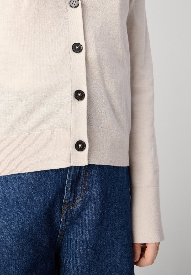 Helles beiges Cardigan mit Rundhalsausschnitt und schwarzen Knöpfen. Gerippte Bündchen und Saum, kombiniert mit blauen Jeans. Glatte Textur.