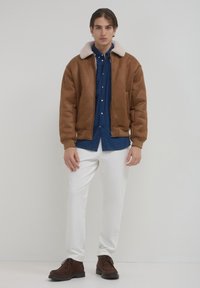 Braune Fake-Leder-Bomberjacke mit kragen aus beige Schafsleder, getragen über einem blauen Hemd mit Knöpfen, kombiniert mit weißen Hosen und braunen Schuhen.