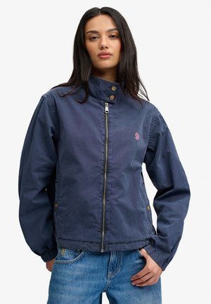 Joven mujer con cabello largo y oscuro que lleva una chaqueta azul marino con cremallera y vaqueros azules, posando con una mano en el bolsillo.