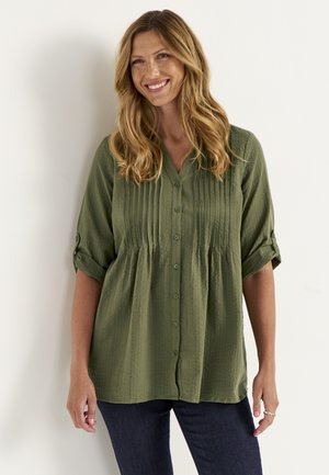 Vrouw met lang golvend haar die lacht, draagt een groene blouse met knopen en opgerolde mouwen en donkerblauwe spijkerbroek, staand tegen een witte achtergrond.