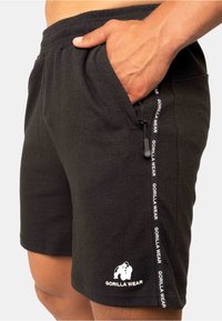 Zwarte sportshorts van gestructureerde stof, met zijzakken en een wit logo met tekst langs de zoom. Ritsdetail op de zijzak.