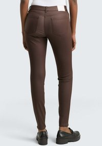 Oseba v urbani krojni rjavih hlačah, črnih loaferjih in belem vrhu, stoji obrnjen/a stran od svetlo sivega ozadja.