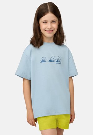 Uśmiechnięta dziewczyna z brązowymi włosami do ramion, ubrana w jasnoniebieski T-shirt z grafiką gór oraz jaskrawożółte krótkie spodenki.