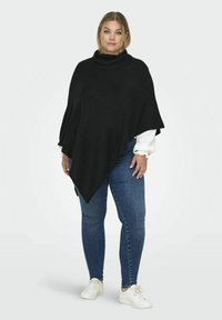 Poncho noir en tricot avec un col montant, présentant un design fluide et un ourlet asymétrique ; porté sur une chemise à manches longues blanche et un jean.