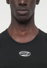 Μαύρο αμάνικο τοπ με εφαρμοστή γραμμή, υφασμάτινη υφή και ασημί λογότυπο κοντά στο λαιμό. Απλός, μινιμαλιστικός σχεδιασμός.