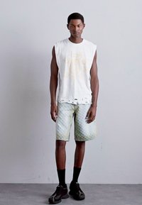 Ærmeløs hvid skjorte med slidte kanter, lysegrønne mønstrede shorts, sorte sokker og sorte sko. Modellen står foran en neutral baggrund.