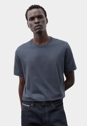 Ung mand med kort krøllet hår iført en ensfarvet mørkegrå T-shirt og mørkeblå jeans mod en lysegrå baggrund.