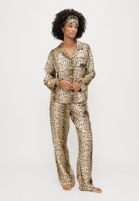 Pijama din satin cu imprimeu de leopard, cu mâneci lungi, pantaloni lejeri și o bandă de cap asortată. Prezintă accente negre și închidere cu nasturi.