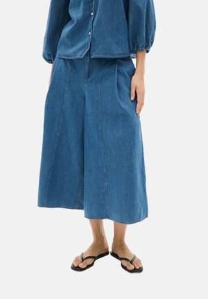 Blaue Denim-Wide-Leg-Culottes mit Falten, hohe Taille und rohem Saum, kombiniert mit schwarzen Flip-Flops. Das Material wirkt strapazierfähig und strukturiert.