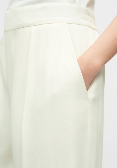 Δεξί χέρι τοποθετημένο στην πλαϊνή τσέπη λευκών παντελονιών με ψηλή μέση και φαρδιά ζώνη.