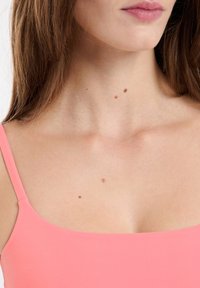 Różowa bezrękawnik z kwadratowym dekoltem. Gładki, elastyczny materiał o jednolitej fakturze. Widoczna skóra z trzema małymi znamionami w pobliżu obojczyka.